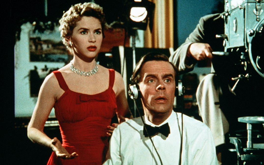 Filmstill aus SIMON AND LAURA: Eine Frau in einem eleganten roten Kleid und ein Mann in einem weißen Hemd mit Fliege stehen neben einer großen Filmkamera und blicken auf das Gefilmte.