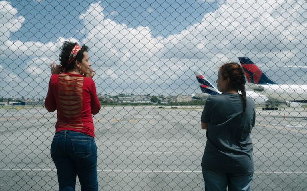 Filmstill aus O ESTRANHO: Zwei Frauen blicken durch einen Maschendrahtzaun auf ein Flugfeld.