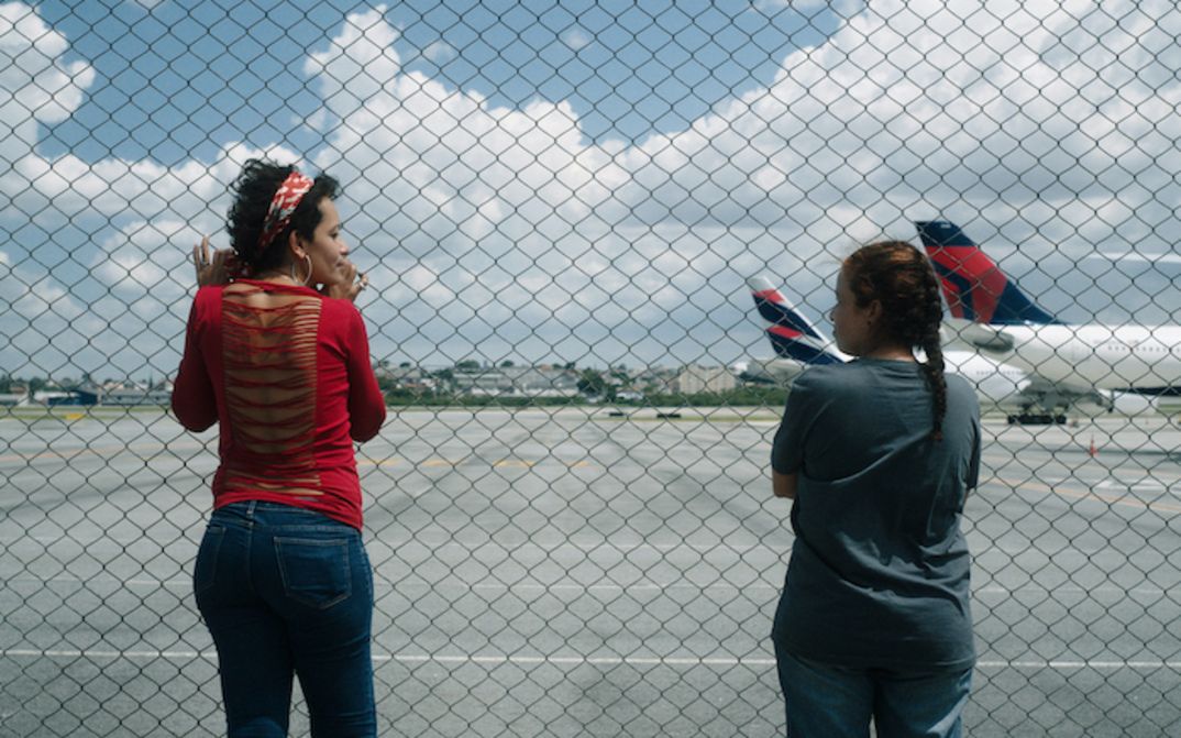 Filmstill aus O ESTRANHO: Zwei Frauen blicken durch einen Maschendrahtzaun auf ein Flugfeld.