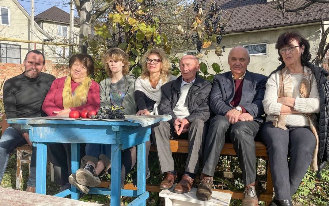 Sieben Leute sitzen nebeneinander auf einer Bank im Garten vor einem Tisch mit Obst, im Hintergrund ein Wohnhaus.