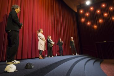 Auf einer großen Bühne sieht man unscharf mehrere Menschen in weitem Abstand stehen. Im Vordergrund des Bildes, scharf, liegen zwei FFP2-Masken.