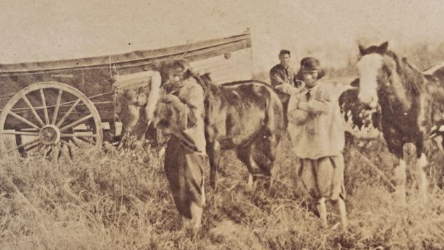 Sepiafarbene Archivaufnahme: Mehrere Personen stehen neben Pferden und einem Wagen auf einem Feld.