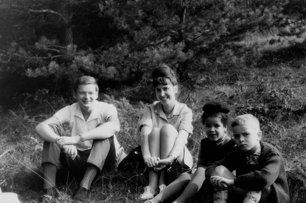 Film still from BECOMING BLACK. A family of four sits on the grass and smiles at the camera. The girl is black, the other family members are white.