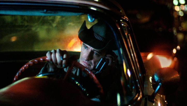 Filmstill aus DER AMERIKANISCHE FREUND: Ein Mann sitz am Lenkrad. Er hat einen Cowboyhut auf und hält ein Telefon ans Ohr. Es ist dunkel und Straßenlichter sind zu sehen.