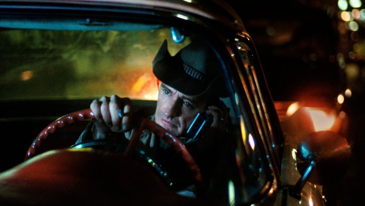 Filmstill from DER AMERIKANISCHE FREUND: A man is sitting at the steering wheel. He has a cowboy hat on and is holding a phone to his ear. It is dark and streetlights can be seen.