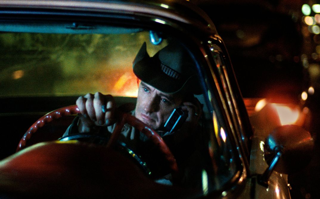 Filmstill from DER AMERIKANISCHE FREUND: A man is sitting at the steering wheel. He has a cowboy hat on and is holding a phone to his ear. It is dark and streetlights can be seen.