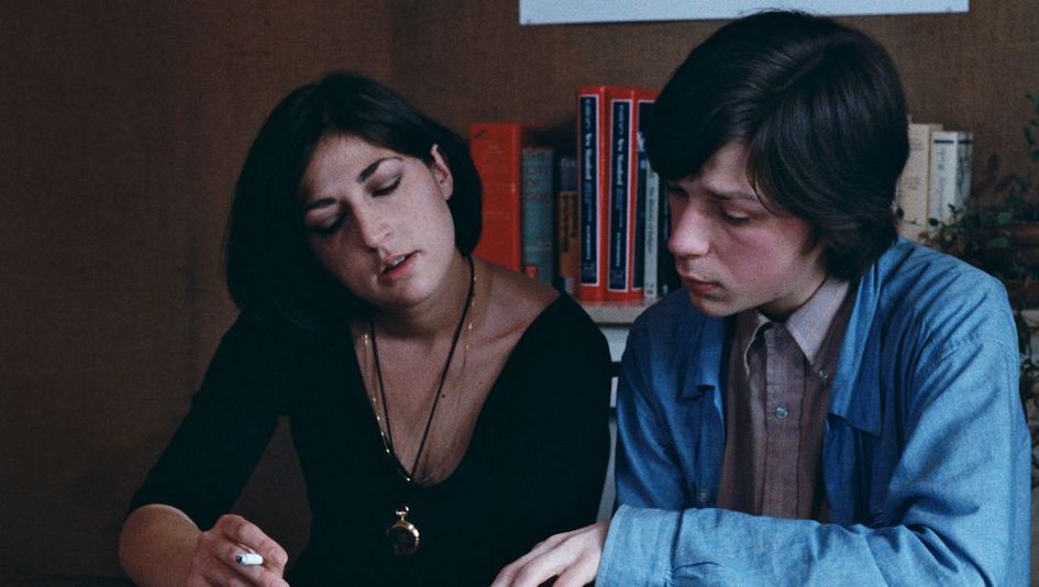 Filmstill aus LES PHOTOS D'ALIX: Zwei junge Menschen betrachten Fotos, die vor ihnen auf dem Tisch liegen. Eine der Personen hält eine Zigarette zwischen den Fingern.