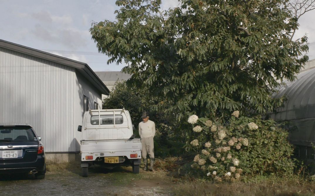 Filmstill aus JAPAN - BIG LAGOON VILLAGE: Ein Mann in Gummistiefeln steht zwischen Bäumen und einem Pick-Up-Wagen.