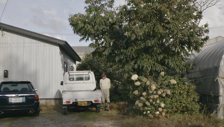 Film still from JAPAN - BIG LAGOON VILLAGE: A man in rubber boots stands between trees and a pick-up truck.