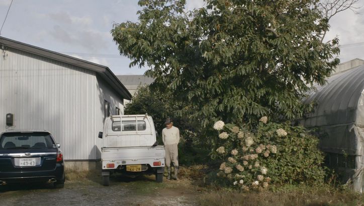 Filmstill aus JAPAN - BIG LAGOON VILLAGE: Ein Mann in Gummistiefeln steht zwischen Bäumen und einem Pick-Up-Wagen.