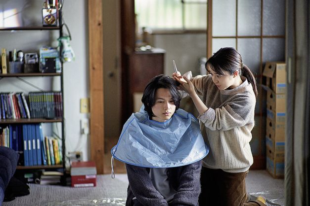 Filmstill aus "Yoake no subete" von Shô Miyake. Zu sehen ist, wie eine Person einer anderen Person zu Hause auf dem Boden die Haare schneidet. 