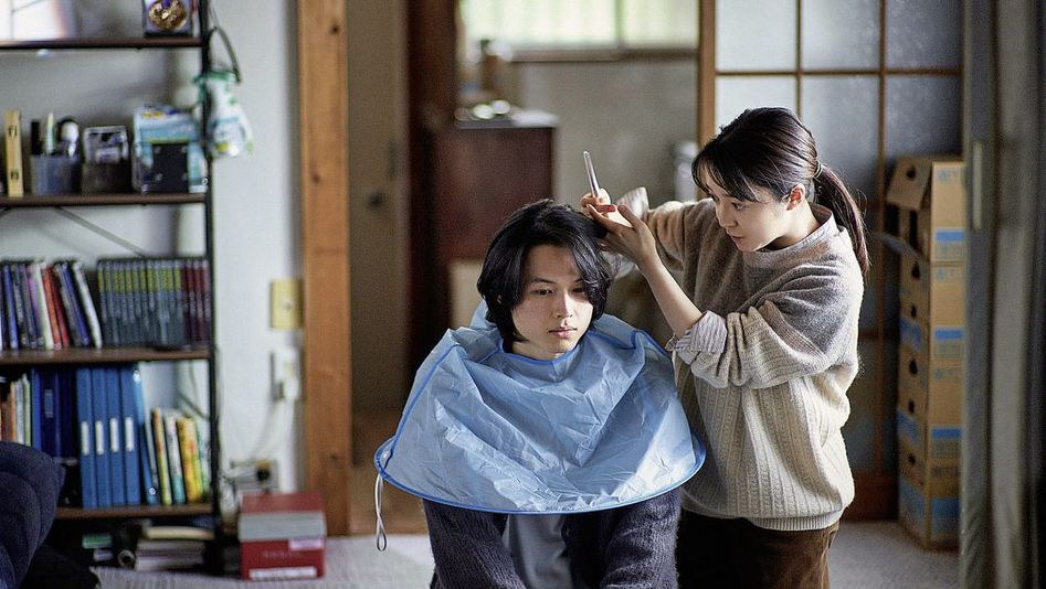 Film still from "Yoake no subete" by Shô Miyake. It shows a person cutting another person's hair on the floor at home.