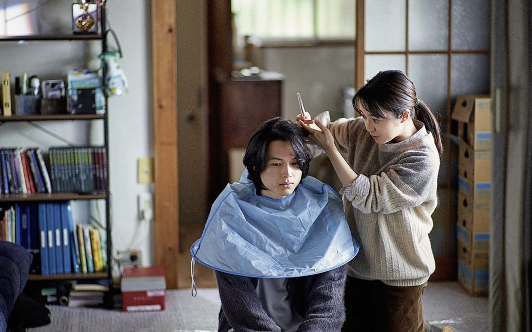 Filmstill aus "Yoake no subete" von Shô Miyake. Zu sehen ist, wie eine Person einer anderen Person zu Hause auf dem Boden die Haare schneidet. 