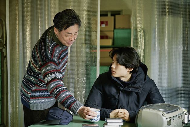 Film still from "Yoake no subete" by Shô Miyake. It shows a young man at a table and an older man placing a cup on the table. The young man looks at the other. 