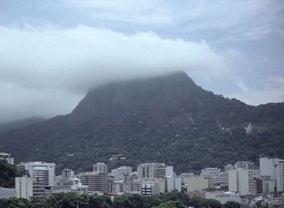 Filmstill aus dem Film „A árvore“ von Ana Vaz. Eine Stadt mit vielen Hochhäusern vor einem Berg, dessen Gipfel von Wolken verdeckt wird.