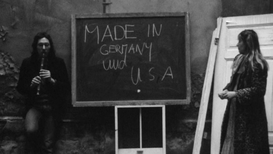 Filmstill aus MADE IN GERMANY AND USA: Ein Mann und eine Frau stehen links und rechts vor einer Wandtafel, auf die der Titel des Films geschrieben steht. Der Mann spielt Flöte.