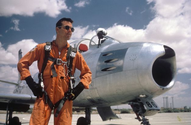 Filmstill aus THE RIGHT STUFF: Ein Pilot steht mit einem orangen Overall und einer Sonnenbrille bekleidet vor einem Flugzeug.
