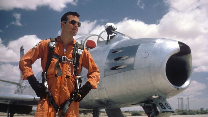 Filmstill aus THE RIGHT STUFF: Ein Pilot steht mit einem orangen Overall und einer Sonnenbrille bekleidet vor einem Flugzeug.