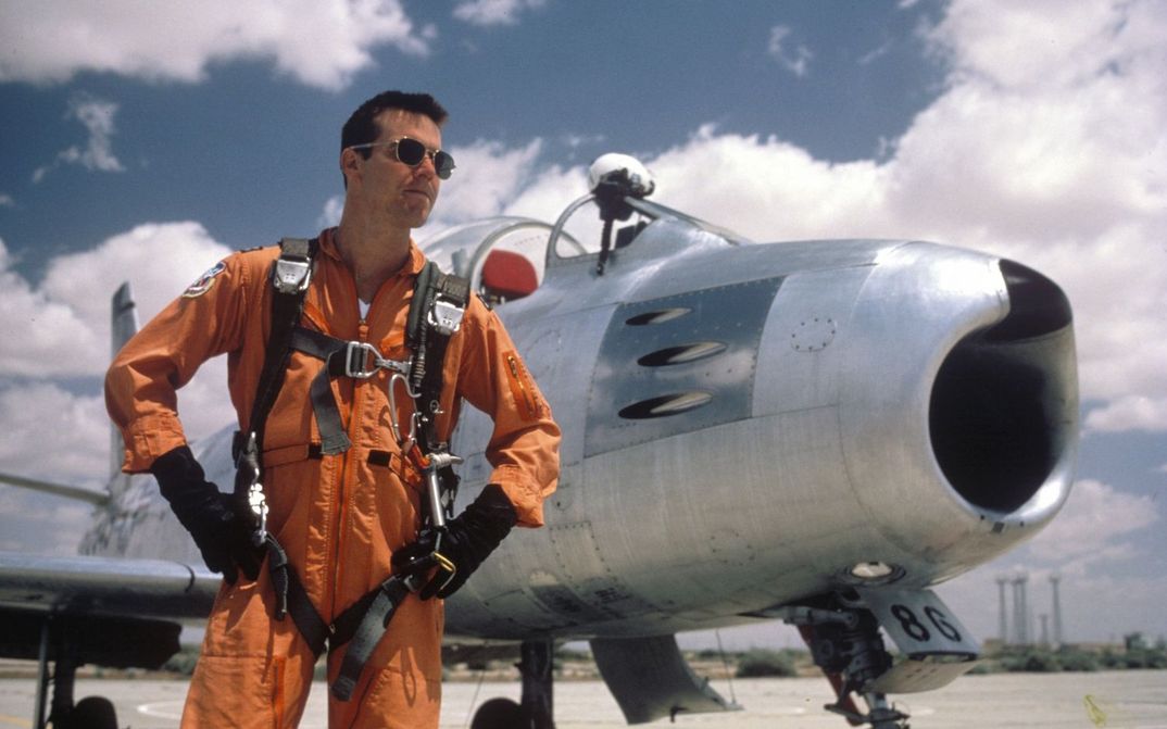 Filmstill aus THE RIGHT STUFF: Ein Pilot steht mit einem orangen Overall und einer Sonnenbrille bekleidet vor einem Flugzeug.
