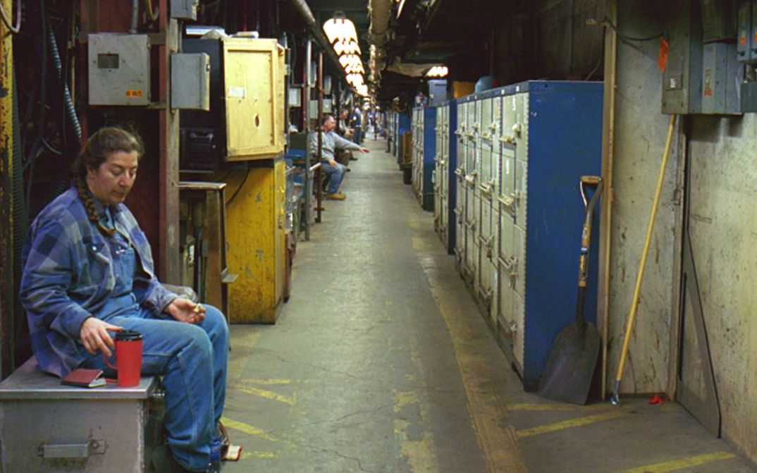 Film still from LUNCH BREAK: A long corridor in a factory. People sit individually in alcoves having lunch.
