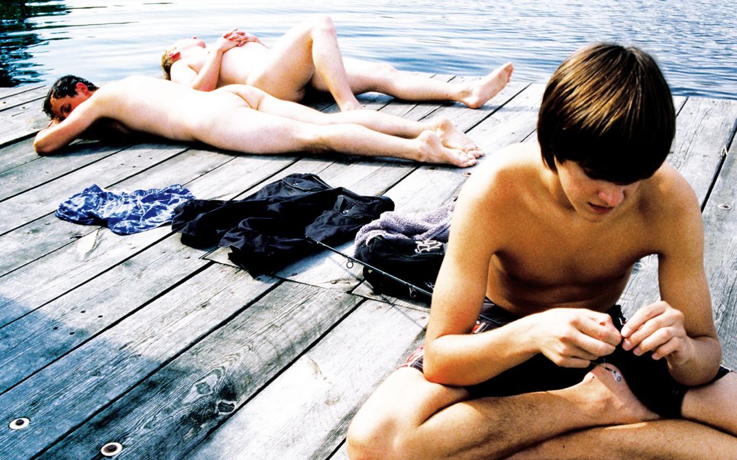 Film still from RÜCKENWIND: Three young men lie and sit on the wooden jetty of a lake.