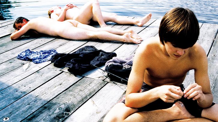 Film still from RÜCKENWIND: Three young men lie and sit on the wooden jetty of a lake. 